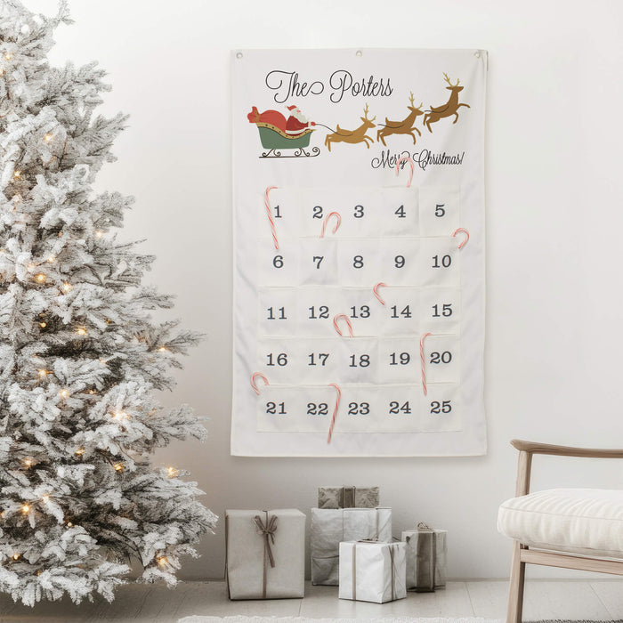 A personalized Christmas advent calendar hanging on a white wall in a decorated living room, next to a flocked Christmas tree and wrapped gifts.