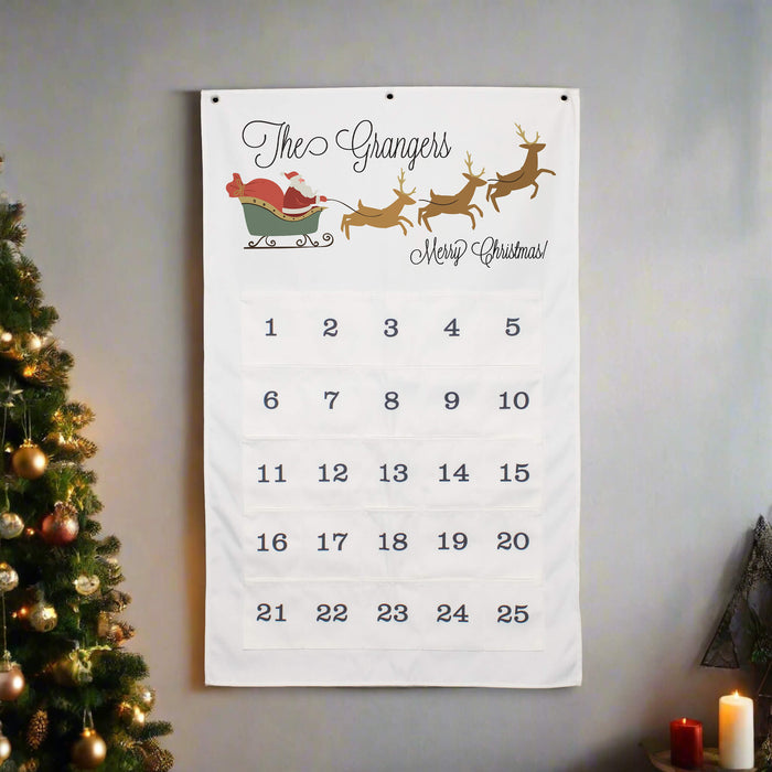 A white Christmas advent calendar hanging on a gray wall, decorated with a festive illustration of Santa Claus in a sleigh and three reindeer. The calendar features 25 numbered pockets for the days of Advent, with 'The Grangers' and 'Merry Christmas!' written at the top.