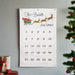 A white fabric wall calendar hanging on a light gray wall, decorated with 'The Burdetts' holiday text, Santa in a sleigh, reindeer, and 'Merry Christmas!' above a grid of numbered pockets 1 through 25. A Christmas tree and wrapped gifts are partially visible at the edges.