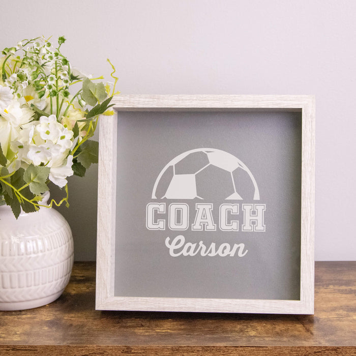 Framed sign with 'COACH Carson' text and soccer ball design on a wooden surface with flowers.