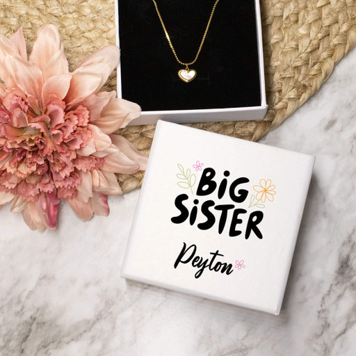 Necklace with heart pendant on a 'Big Sister' box, surrounded by flowers on a marble surface.