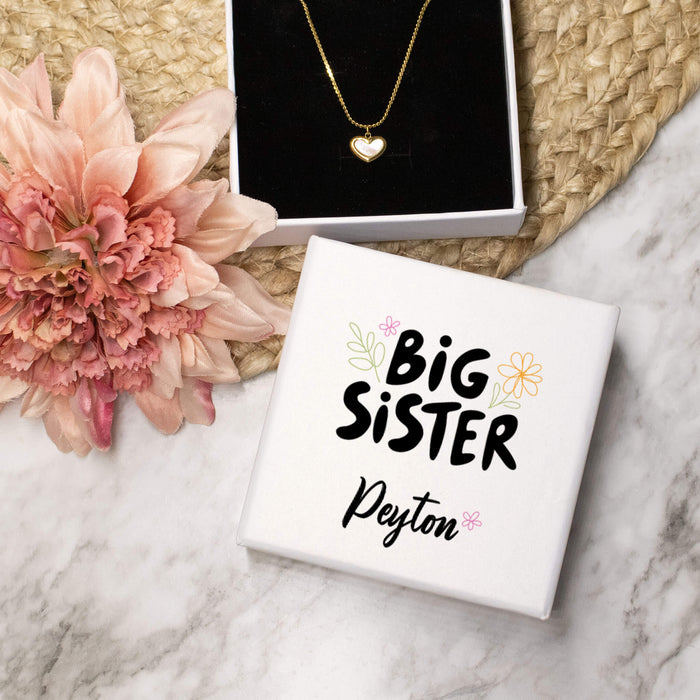 Necklace with heart pendant on a 'Big Sister' box, surrounded by flowers on a marble surface.
