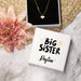 Necklace with heart pendant on a 'Big Sister' box, surrounded by flowers on a marble surface.