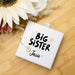 Necklace with 'Big Sister' on a white box, surrounded by flowers on a wooden surface