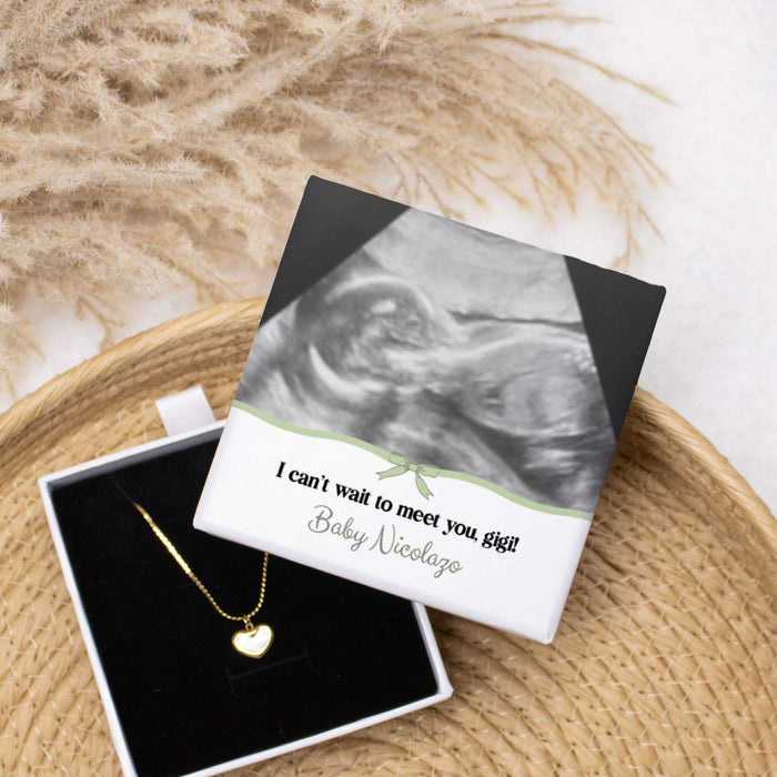 Gold heart necklace in a box that has an ultrasound picture printed on it laid on top of a basket and pampas grass