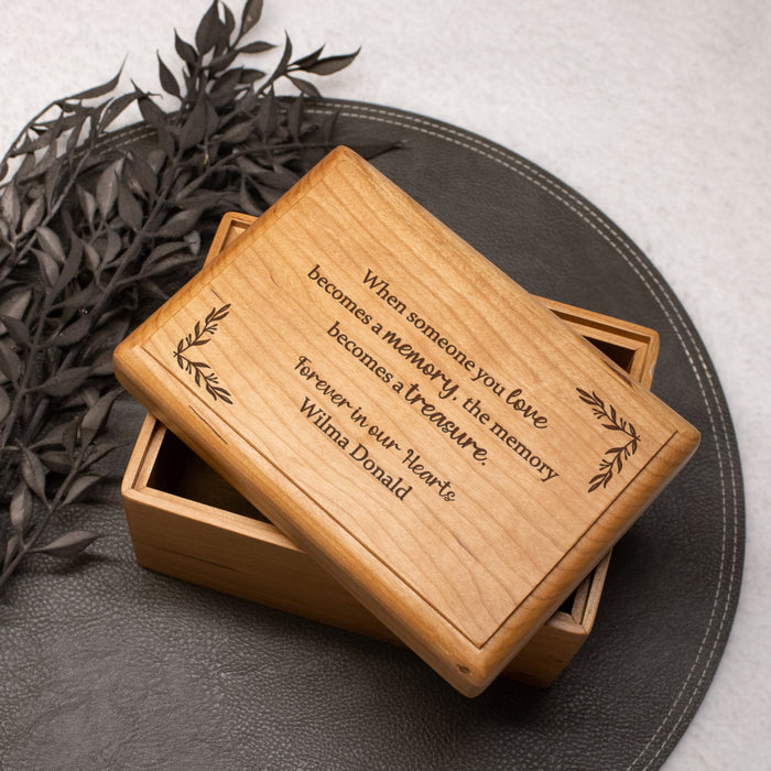Wooden memorial keepsake box with engraved custom text on a dark surface with leaves.