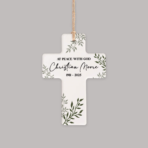 A white cross-shaped memorial ornament with green leaf illustrations and black personalized text reading 'AT PEACE WITH GOD Christian Moore 1981 - 2025', hanging from a natural jute string against a solid gray background.
