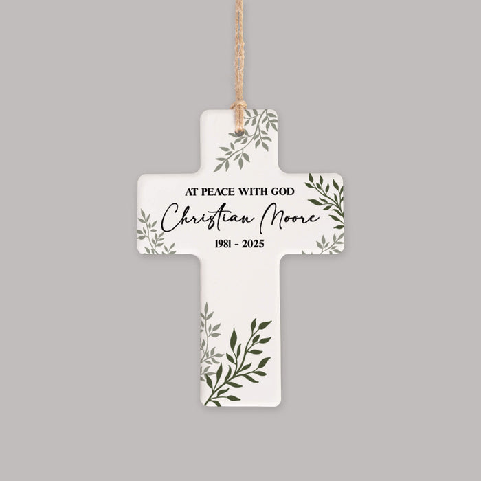 A white cross-shaped memorial ornament with green leaf illustrations and black personalized text reading 'AT PEACE WITH GOD Christian Moore 1981 - 2025', hanging from a natural jute string against a solid gray background.