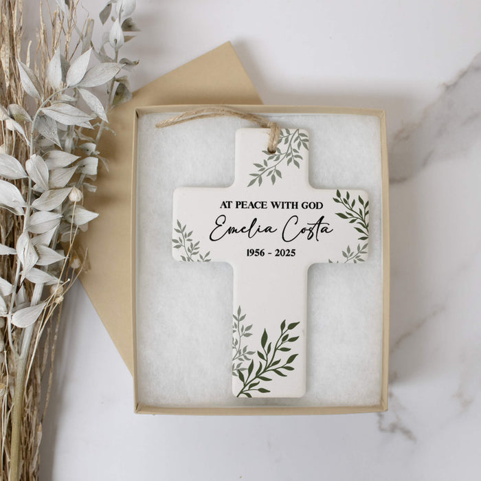 A personalized ceramic cross ornament keepsake in a light beige box with white cushioning, styled on a white marble surface with dried white leaves.