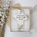A personalized ceramic cross ornament keepsake in a light beige box with white cushioning, styled on a white marble surface with dried white leaves.