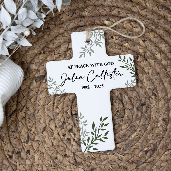 A white cross hanging ornament with green leaf illustrations and personalized text, displayed on a woven brown mat with white dried flowers as props.