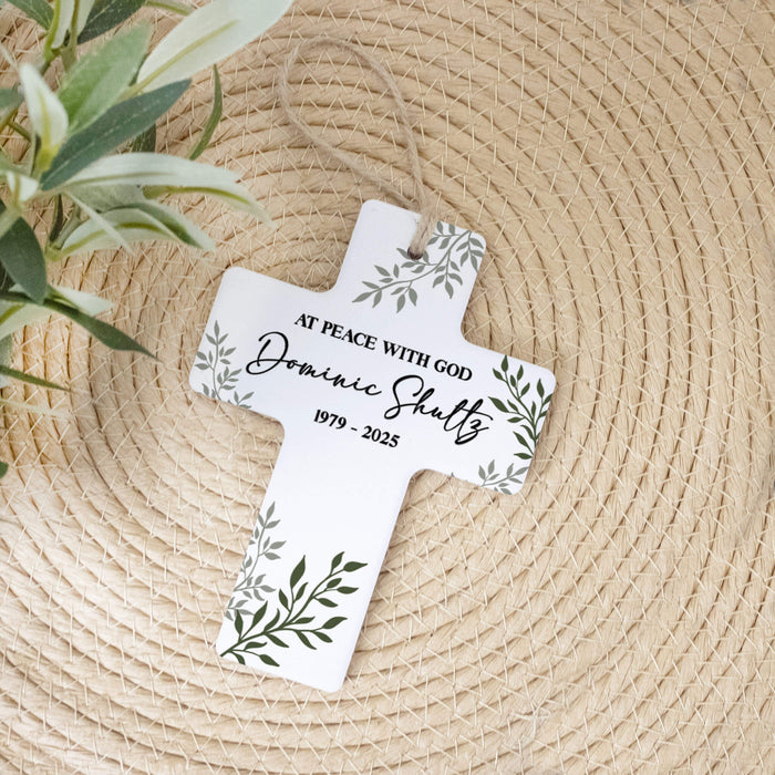 A personalized white cross hanging ornament with green leaf illustrations and black text, displayed on a woven straw mat with a plant in the corner.