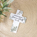 A personalized white cross hanging ornament with green leaf illustrations and black text, displayed on a woven straw mat with a plant in the corner.