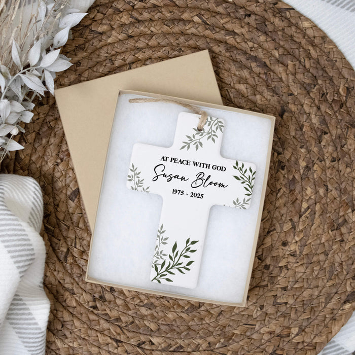 Personalized cross memorial ornament with green leaf illustrations and the text 'AT PEACE WITH GOD Susan Bloom 1975 - 2025', displayed in a gift box on a woven placemat.