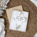 Personalized cross memorial ornament with green leaf illustrations and the text 'AT PEACE WITH GOD Susan Bloom 1975 - 2025', displayed in a gift box on a woven placemat.