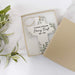 A personalized ceramic cross keepsake ornament with a hanging tag, displayed inside an open gift box on a white marble surface with green leaves at the side.