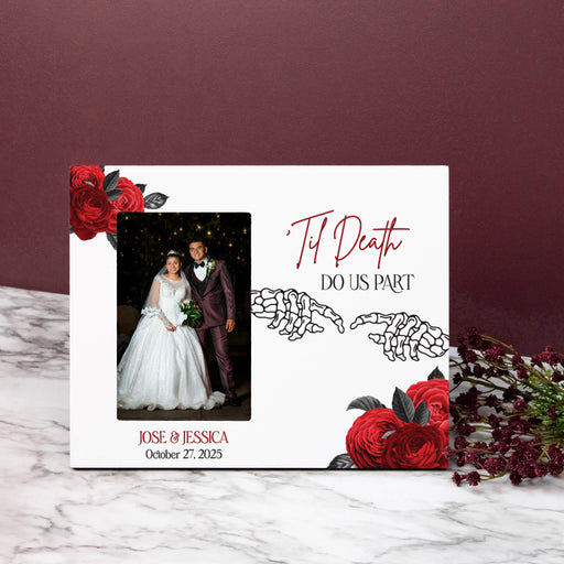 A personalized white photo frame styled on a white marble surface against a dark burgundy background. The plaque features red roses, skeleton hands, and the text 'Til Death Do Us Part' along with a custom wedding photo of a bride and groom.