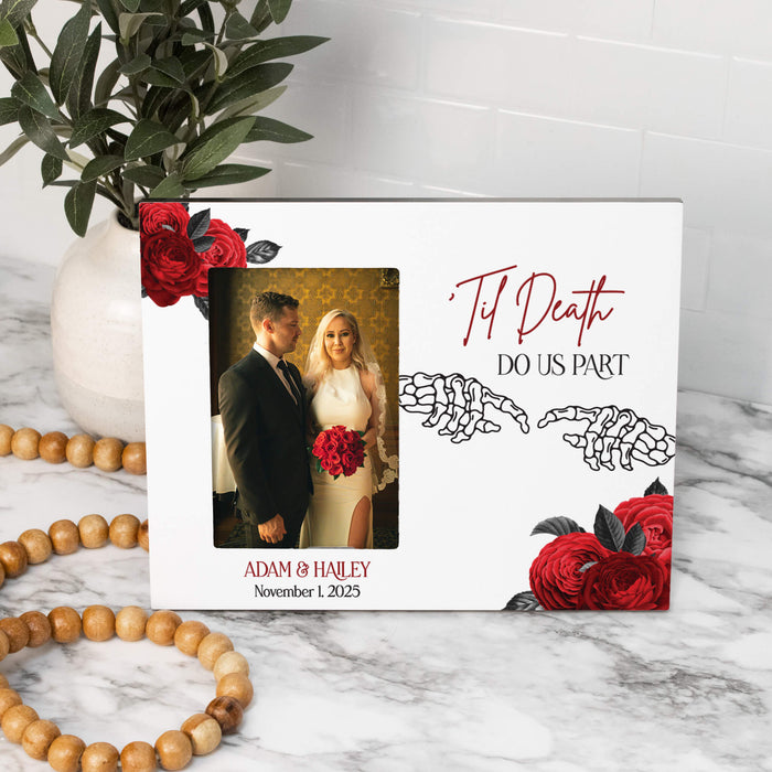 A white personalized photo frame with red roses, skeleton hands, and the text 'Til Death DO US PART' and 'ADAM & HALEY November 1, 2025', styled on a marble surface with a vase of greenery and wooden beads nearby.
