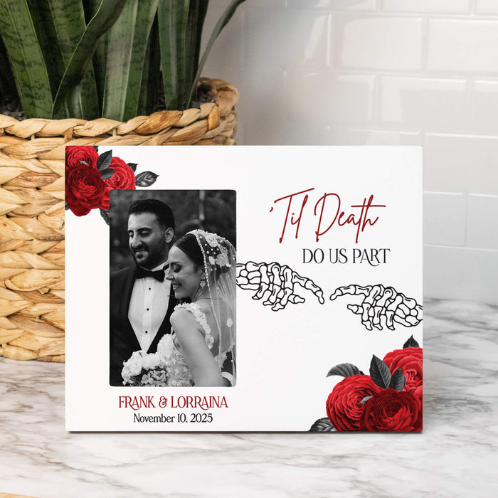 A custom skeleton wedding photo frame displayed on a marble surface next to a wicker basket with a plant. The white picture frame features a black-and-white photo of a bride and groom, red roses, skeleton hands reaching together, and the text 'Til Death DO US PART' and 'FRANK & LORRAINA November 10, 2025'.