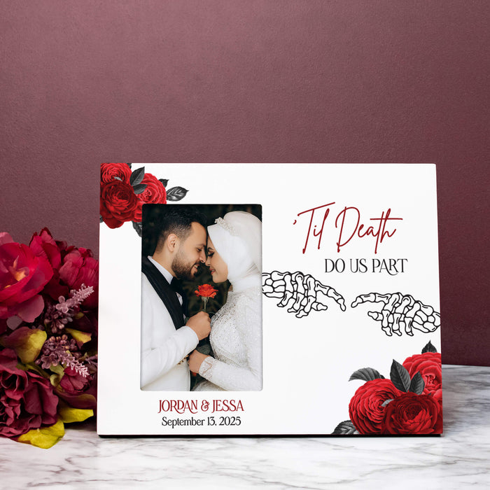 A white personalized photo frame with red rose illustrations and skeleton hands, featuring a custom wedding photo of a couple and the text 'Til Death Do Us Part' and 'Jordan & Jessa September 13, 2025'. The frame is styled on a marble surface next to a bouquet of red flowers against a dark mauve background.
