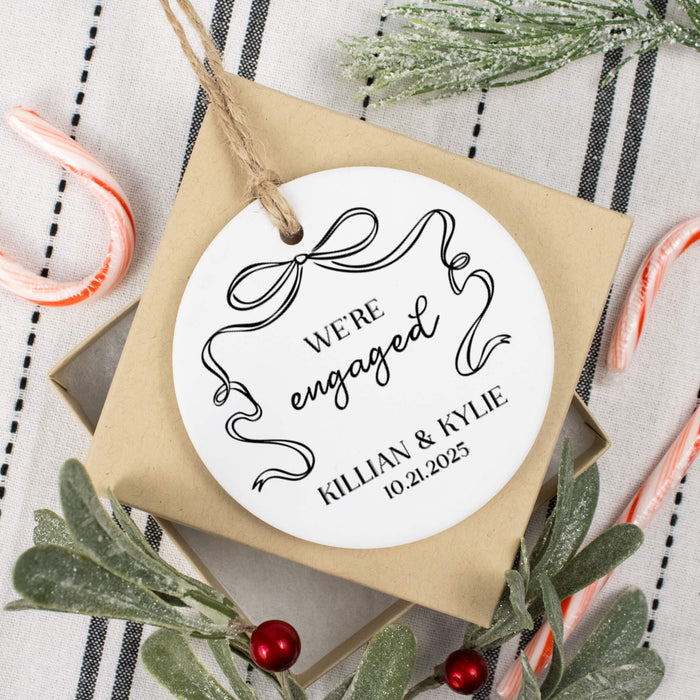 Round white ornament with 'We're Engaged' text on a square brown gift box, surrounded by Christmas decorations.