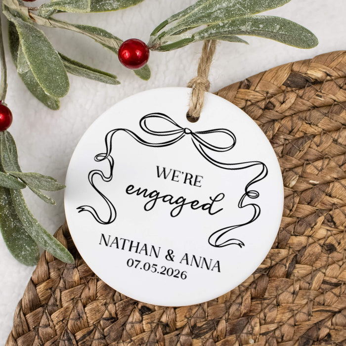 Round white ornament with 'We're engaged' text and personalization on a woven surface with greenery.