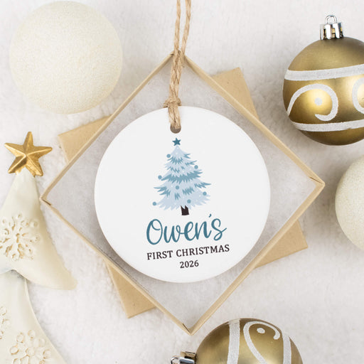 A personalized round white Christmas ornament hanging from a string, displayed in a gift box. The ornament features a light blue Christmas tree and the text 'Owen's FIRST CHRISTMAS 2026'.