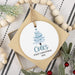 A personalized round ceramic Christmas ornament hanging from a string, featuring a light blue Christmas tree and the text 'Cole's FIRST CHRISTMAS 2026'. The ornament is displayed on a square gift box with a neutral striped fabric background and holiday decorations.