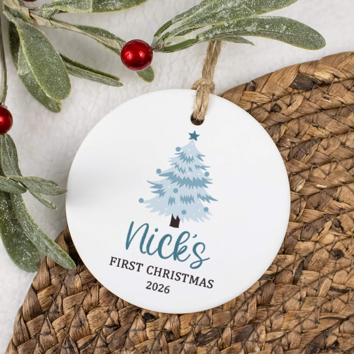 A round white ceramic Christmas ornament with a personalized print of a blue Christmas tree and the text 'Nick's FIRST CHRISTMAS 2026', hanging from a twine string. The ornament is styled on a brown woven placemat with green frosted branches and red berries in the background.