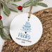 A round white ceramic Christmas ornament with a personalized print of a blue Christmas tree and the text 'Nick's FIRST CHRISTMAS 2026', hanging from a twine string. The ornament is styled on a brown woven placemat with green frosted branches and red berries in the background.