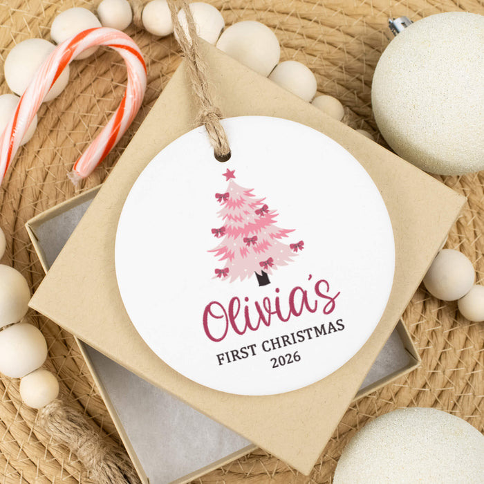 A round white personalized Christmas ornament hanging from a string, featuring a pink Christmas tree and the text 'Olivia's FIRST CHRISTMAS 2026'. The ornament is displayed on a square gift box surrounded by holiday decorations including candy canes, white beads, and a white ornament on a woven jute background.