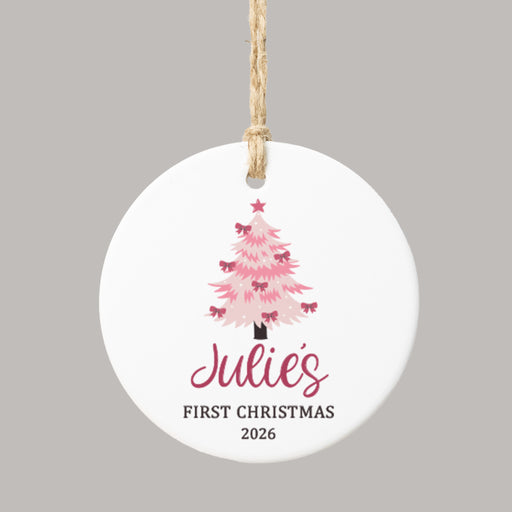 A round white ceramic Christmas ornament with a hanging twine loop, featuring a pink Christmas tree, the personalized name 'Julie's' in dark pink script, and the text 'FIRST CHRISTMAS 2026' below, shown on a solid gray background.