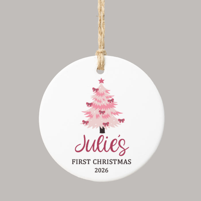 A round white ceramic Christmas ornament with a hanging twine loop, featuring a pink Christmas tree, the personalized name 'Julie's' in dark pink script, and the text 'FIRST CHRISTMAS 2026' below, shown on a solid gray background.