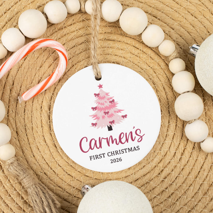 A personalized round white Christmas ornament hanging from a jute string, featuring a pink illustrated Christmas tree and the text 'Carmen's FIRST CHRISTMAS 2026'. The ornament is styled on a woven straw mat with wooden beads, a candy cane, and other holiday decorations.
