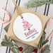A round white Christmas ornament with a pink tree and the text 'Bella's FIRST CHRISTMAS 2026' is displayed on a gift box. The scene is styled with candy canes, green foliage, and red berries on a striped fabric background.