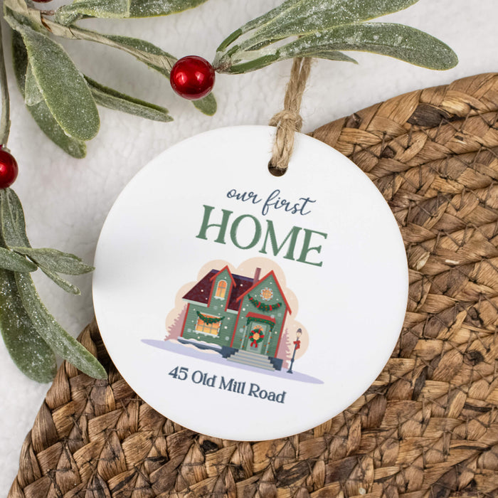 A round white ceramic Christmas ornament hanging from a twine loop, printed with the phrase "our first HOME" and a small illustrated house, with the address "45 Old Mill Road" below. The ornament is styled on a woven brown placemat with green frosted branches and red berries in the background.