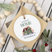 A square gift box with a round white hanging ornament printed with the phrase 'our first HOME' and a small illustrated house, plus the address '330 McCallister St.' The box is styled on a striped fabric with wooden beads and holiday greenery.