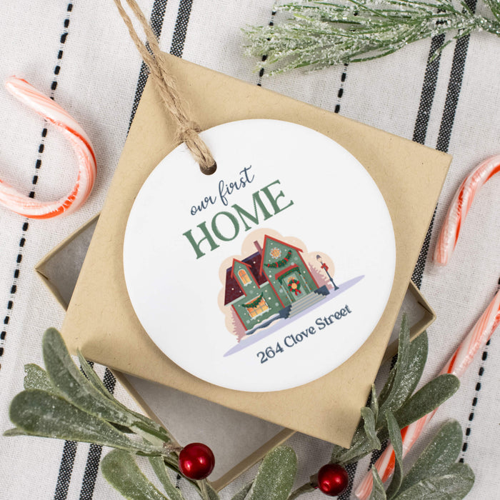 A round white Christmas ornament with a personalized illustration of a house and the text 'our first HOME 264 Clover Street' on top of a gift box. The ornament is styled in a holiday flat-lay arrangement with candy canes, green foliage, and red berries on a striped fabric background.