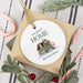 A round white Christmas ornament with a personalized illustration of a house and the text 'our first HOME 264 Clover Street' on top of a gift box. The ornament is styled in a holiday flat-lay arrangement with candy canes, green foliage, and red berries on a striped fabric background.