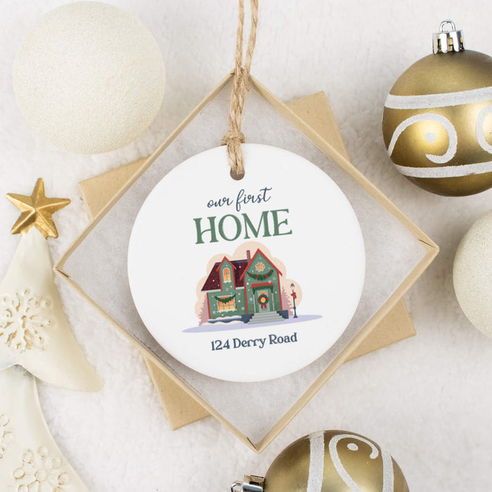 A personalized round ceramic Christmas ornament hanging from a twine string, displayed in a diamond-shaped box with holiday decorations around it. The ornament features the text 'our first HOME' and the address '124 Derry Road' above a colorful illustration of a decorated house.