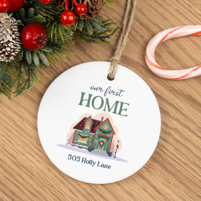 A round white ceramic Christmas ornament hanging on a wooden table, printed with the phrase 'our first HOME' and the address '505 Holly Lane' above a small illustrated house. Festive greenery with red berries and a candy cane are partially visible around it.
