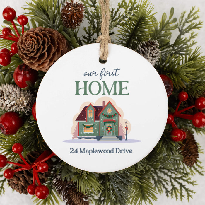 A round white ceramic Christmas ornament hanging from a twine loop, printed with the phrase 'our first HOME' above a small illustrated house and the address '24 Maplewood Drive.' The ornament is centered against a festive wreath made of green pine branches, red berries, and pinecones.