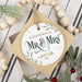 personalized Christmas ornament placed on top of its gift box next to white beads and holly branches on a black and white striped background