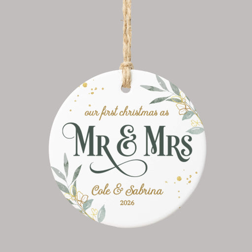 A round white ceramic Christmas ornament with a hanging loop, featuring personalized text that reads 'our first christmas as Mr & Mrs Cole & Sabrina 2026' in dark green and gold lettering, decorated with gold confetti and green leaf illustrations, shown on a solid gray background.