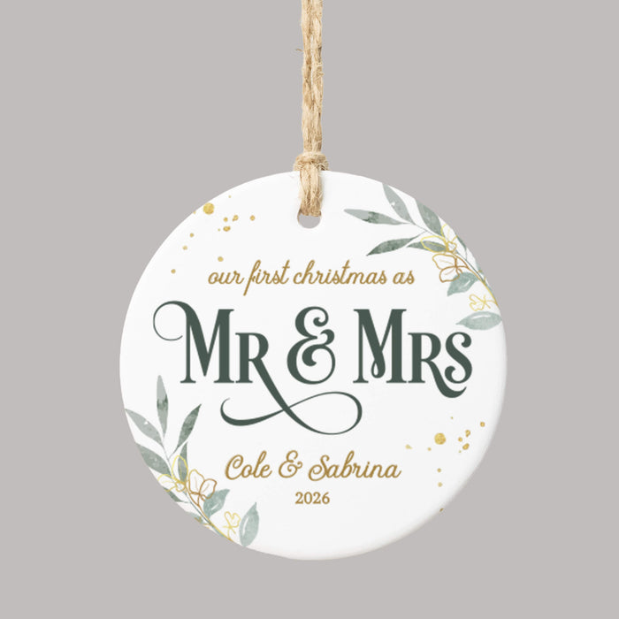 A round white ceramic Christmas ornament with a hanging loop, featuring personalized text that reads 'our first christmas as Mr & Mrs Cole & Sabrina 2026' in dark green and gold lettering, decorated with gold confetti and green leaf illustrations, shown on a solid gray background.
