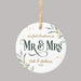 A round white ceramic Christmas ornament with a hanging loop, featuring personalized text that reads 'our first christmas as Mr & Mrs Cole & Sabrina 2026' in dark green and gold lettering, decorated with gold confetti and green leaf illustrations, shown on a solid gray background.