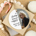 A square paper ornament package containing a round hanging memorial tag with a photo of an older woman and the text 'In Loving Memory Sheryl Lolli 1958 - 2025.' The ornament is styled on a woven mat with candy canes, white beads, and a gift box around it.