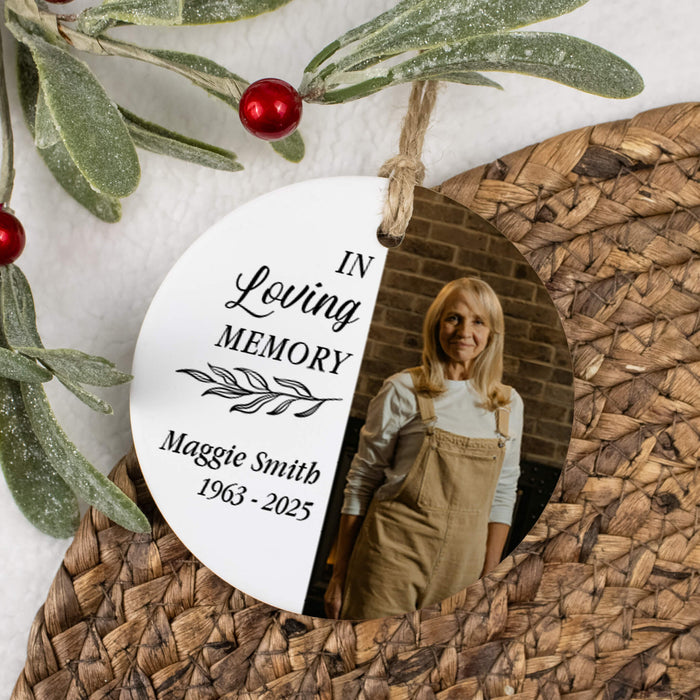 A personalized oval memorial hanging ornament with a photo of a woman and the text 'In Loving Memory Maggie Smith 1963 - 2025', styled on a woven placemat with greenery and red berries.