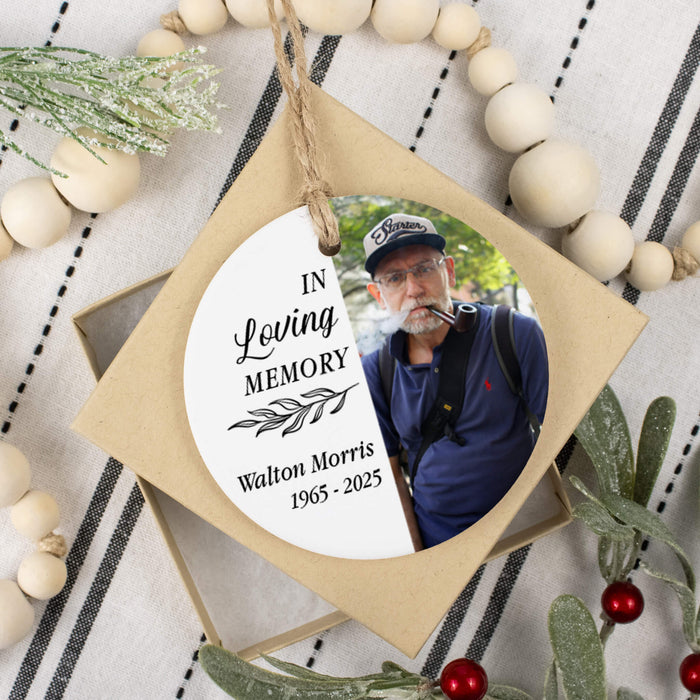 A circle memorial ornament hangs from a string over a small gift box. The ornament features a circular printed photo of a man and memorial text reading 'In Loving Memory' and 'Walton Morris 1965 - 2025.