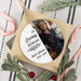 A square gift box on a striped fabric surface, wrapped with a round hanging memorial tag that reads 'In Loving Memory Barb Allister 1946-2025' and includes a printed photo of an older woman. Candy canes and greenery are arranged around the box.
